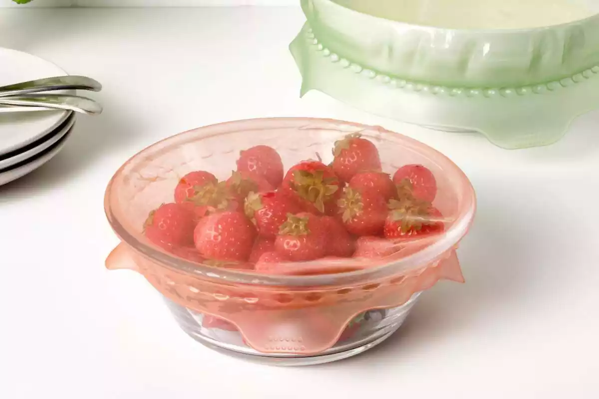 Bol de cristal lleno de fresas con una tapa roja transparente de silicona, junto a fragmento de bol con misma tapa en verda y tres platos blancos apilados con cubiertos