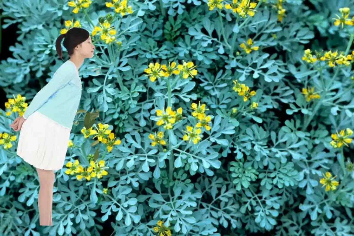 Una mujer con una blusa azul y falda blanca se inclina hacia adelante con los ojos cerrados, rodeada de un fondo de plantas verdes con flores amarillas.